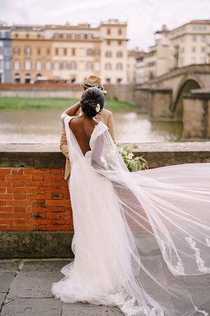 Multiracial wedding couple. Wedding in Florence, Italy. African-American bride and Caucasian groom stand embracing on the embankment of the Arno River, overlooking the city and bridges.の写真素材