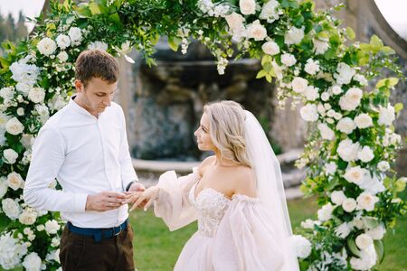 The groom puts the ring on the brides finger. Wedding at an old winery villa in Tuscany, Italy. Round wedding arch decorated with white flowers and greenery in front of an ancient Italian architectureの写真素材