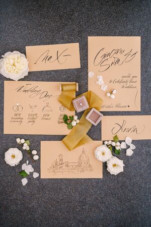 Wedding rings in a box, invitation cards, silk ribbon, white roses on a black background. Overhead...