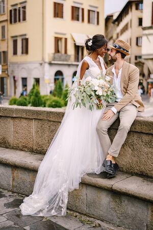 Interracial wedding couple. Wedding in Florence, Italy. African-American bride in a white dress with a long veil and a bouquet, and Caucasian groom in a sandy jacket and straw hat.の写真素材