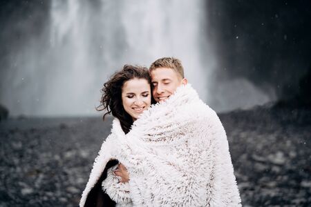 Wedding couple near Skogafoss waterfall. The bride and groom covered with a woolen blanket, where hugging. Snow is falling, falling snowflakes.の写真素材