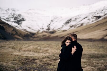 Destination Iceland wedding. Wedding couple on a background of snowy mountains. The bride and groom in black coats are hugging in a field of moss and yellow grass.の写真素材
