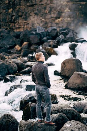 A guy in a black knitted sweater stands on rocks near Ehsaraurfoss Falls, Ehsarau River, National Park, Tingvedlir, Syudurland Region, Iceland.の写真素材