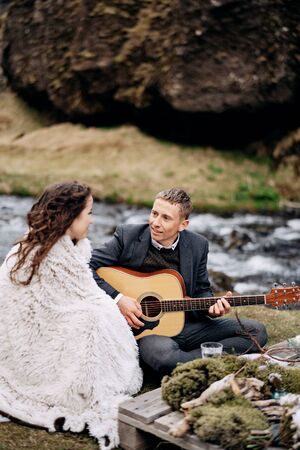 A wedding couple is sitting on the shore of a mountain river, at a table for a wedding dinner. The groom plays and sings for the bride. Destination Iceland wedding, near Kvernufoss waterfall.の写真素材