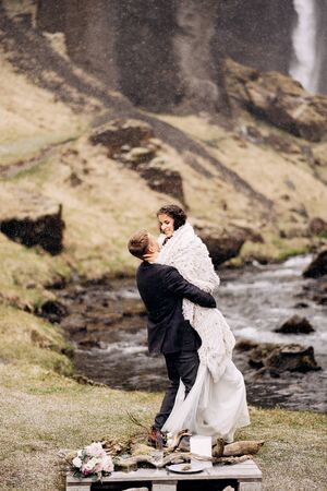 Destination Iceland wedding, near Kvernufoss waterfall. Wedding couple on the shore of a mountain river. The groom wears the bride in a woolen blanket in his hands.の写真素材