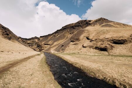 A fast mountain river flows among a field of yellow dry grass, against a backdrop of small mountains in Iceland, against a blue sky with white clouds.の写真素材