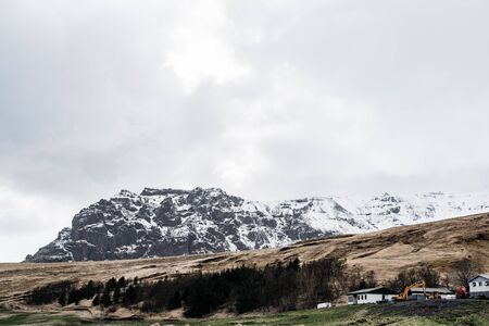 White houses and a construction excavator near the forest on a hill, against the backdrop of a rocky snow-capped mountain in Iceland.の写真素材