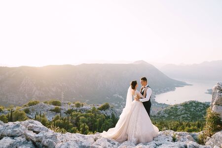 A wedding couple stands on top of a mountain with panoramic views of the Bay of Kotor, at sunset. The groom gently hugs the bride. Fine-art destination wedding photo in Montenegro, Mount Lovchen.の写真素材