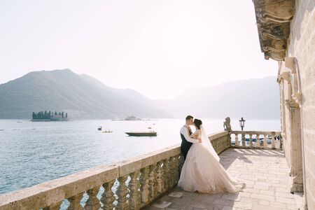 A wedding couple stands on a large terrace with stone columns and statues of lions, overlooking the...