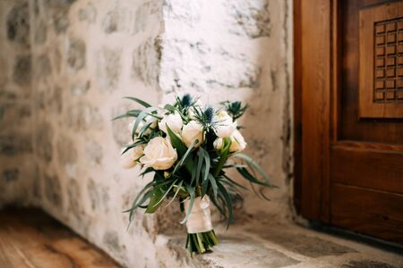 bridal bouquet of pink and white roses, tulips, branches of eucalyptus tree, eryngium, scabiosa and white ribbons on the floor near wooden door and stone wall. High quality photoの写真素材