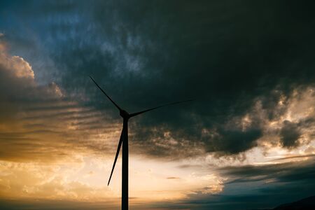 The silhouette of a high industrial wind turbine against the sunset sky with clouds in the evening.の写真素材
