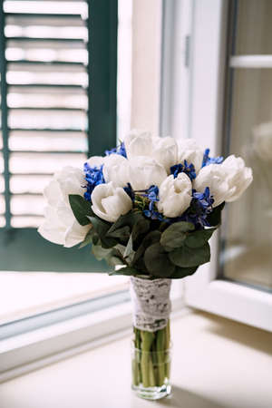 bridal bouquet of white tulips, delphinium, salal, limonium and lace ribbons in glass vase on the windowsillの写真素材