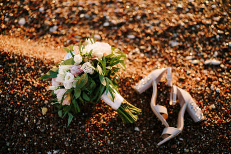 bridal bouquet of white and pink roses, peonies, hortense branches of eucalypt tree, eustoma white ribbons and the shoes of the bride on a pebble beachの写真素材