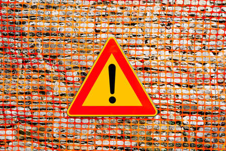 An exclamation point on a yellow background, in a red triangular sign. The texture of the yellow plastic mesh fence on the construction site.の写真素材