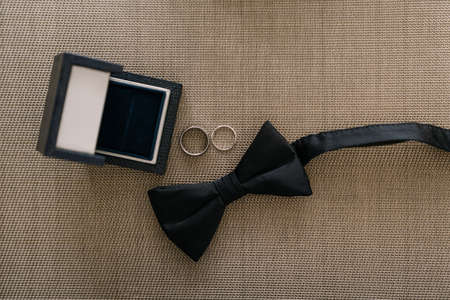 Black bow tie for the groom and two wedding rings with a box on a gray background.