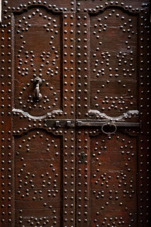Brown wooden doors with many metal rivets and carvings with a long bolt and door knocker.