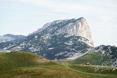 Tourists in a red car, camped on the grass, against the backdrop of a rocky mountain near the Sedlo Pass, in the national park of Durmitor, in northern Montenegro. High quality photoの写真素材