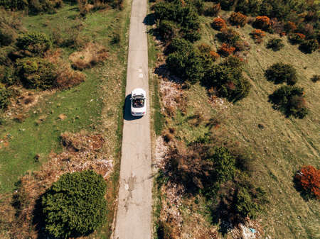 A white convertible drives through the woods, along a paved road, among tall trees. Aerial top view.の写真素材