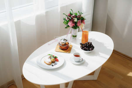 White table inside the room near the window with a bouquet of flowers and breakfast in the morning...