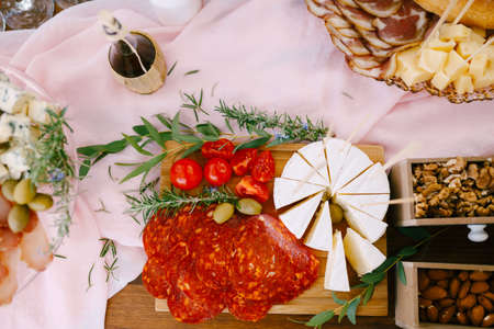Top view of a cutting board with chorizo sausage, brie and tomatoes near wooden boxes of nuts and a plate of ham and hard cheese.の写真素材