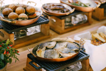 Fried fish on a copper frying pan on a stove against the background of mini burgers in the kitchen...