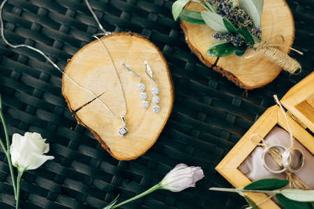 Earrings and a chain with a silver pendant on a wooden stand on a table with a box with wedding rings and a bridal bouquet.の写真素材