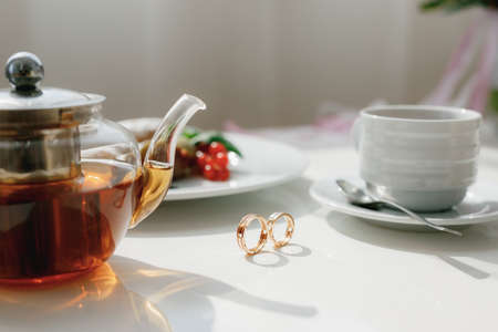 Wedding gold rings on a table with a full teapot and a cup with a spoon on a saucer.の写真素材