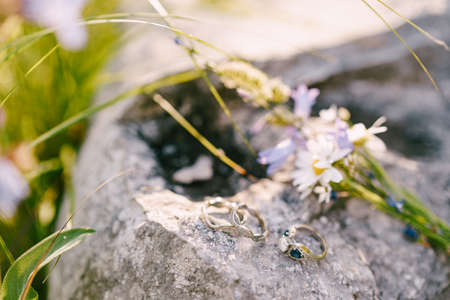 Close-up of wedding rings and an engagement ring on a stone with a bouquet of wildflowers.の写真素材