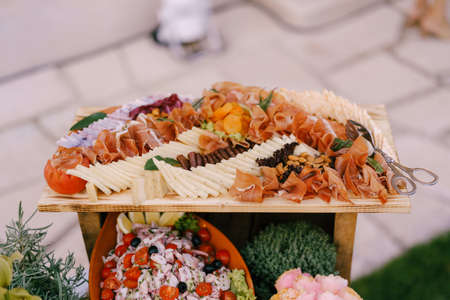 Wooden plank with cheese platter, prosciutto, dried apricots and almonds with salad in a platter.