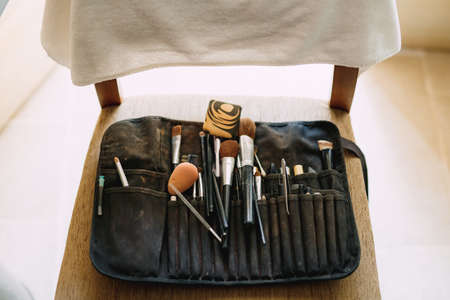 Black work beautician cosmetic bag with makeup brushes, sponge and eyebrow tweezers on wooden chair.の写真素材
