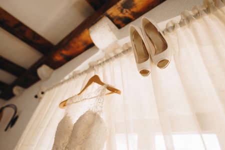The brides wedding dress on a hanger and white shoes on the background of closed curtains on the cornice.の写真素材