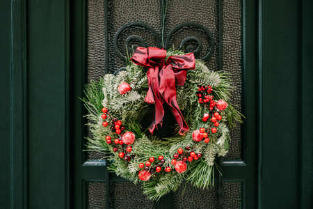 A Christmas wreath of fir and pine branches, with a red bow and berries, hangs on a green wooden...