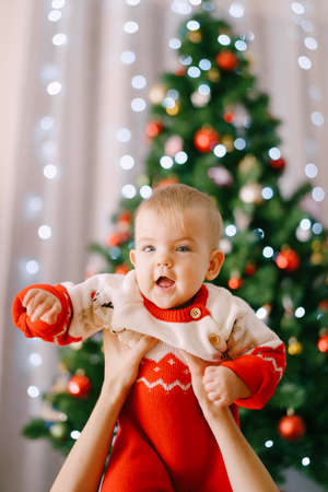 Toddler in a red-and-white Christmas bodysuit in her mothers arms in front of a Christmas treeの写真素材