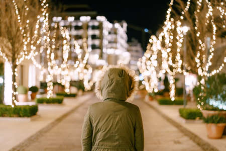 A girl in a hood with fur stands on a street decorated for Christmas in the city.