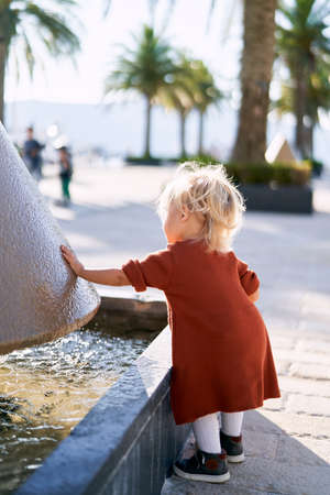 A cute toddler in a terracotta dress and white tights is plaing by the fountain on a sunny dayの写真素材