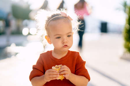 Cute baby girl is eating a pie by the fountain in the parkの写真素材