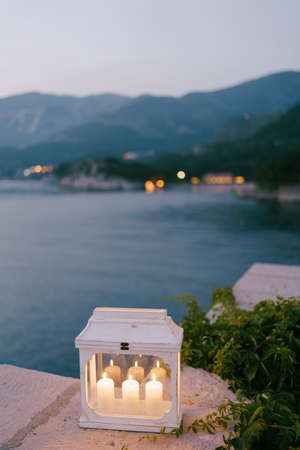 A candlestick-lantern with three lighted candles by ivy branches on a stone border against the background of mountains and water.の写真素材