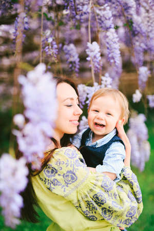 Cute red-haired baby boy laughing in his mothers arms under a wysteria treeの写真素材