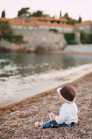 A blue-eyed little boy in a cute hat is sitting on the seashore against the background of the Sveti Stefan island, back viewの写真素材