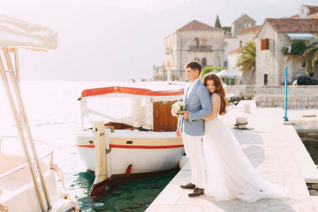 The bride hugs the groom from behind on the pier near the old town of Perast, next to them is a...