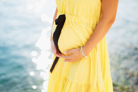 A pregnant woman in a cute bright yellow dress stands by the sea and holds her belly with her hands, close-upの写真素材