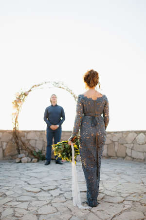 A bride with a bouquet in her hand goes to the groom who is waiting for her at the wedding archの写真素材
