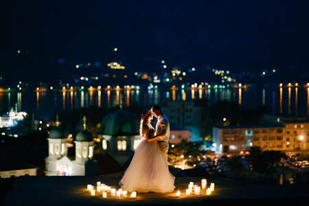 The bride and groom embrace at sunset on the observation deck above the old town of Kotor, behind them the evening harbor in lightsの写真素材