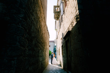 The bride and groom hold hands on a beautiful old street of Perast in a narrow dark passage between housesの写真素材