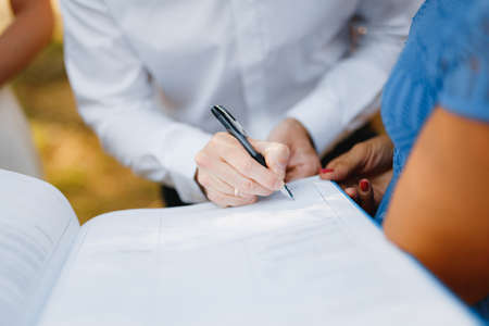 The groom signs the wedding certificate during the wedding ceremony, the bride stands near him, close-up.の写真素材