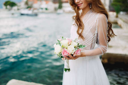The bride holds a bouquet of roses in her hands and stands on the pier near the old town of Perastの写真素材