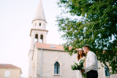 The bride and groom walk through the old town of Budva, the groom gently hugs the bride and holds a bouquet in his handの写真素材