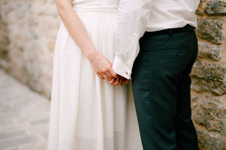 The bride and groom stand near a stone wall and touchingly hold each others hands, close-upの写真素材