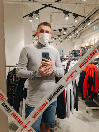 Budva, Montenegro - 01 august 2020: A man wearing a medical mask takes a selfie on his phone in the mirror of closed fitting rooms, in connection with the coronavirus pandemic in 2020.のeditorial素材