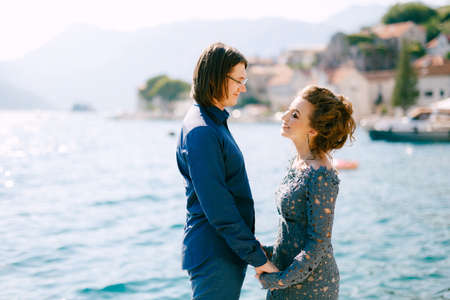 The bride in a stylish grey wedding dress and the groom stand holding hands on the seashore near the...